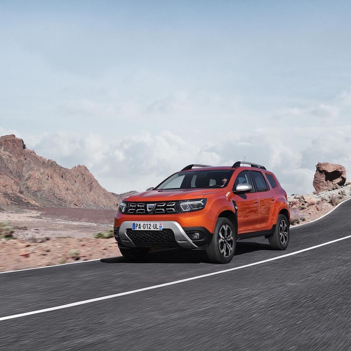 Orange Dacia Duster SUV driving on an asphalt road with a desert mountain landscape and a cloudy blue sky background.