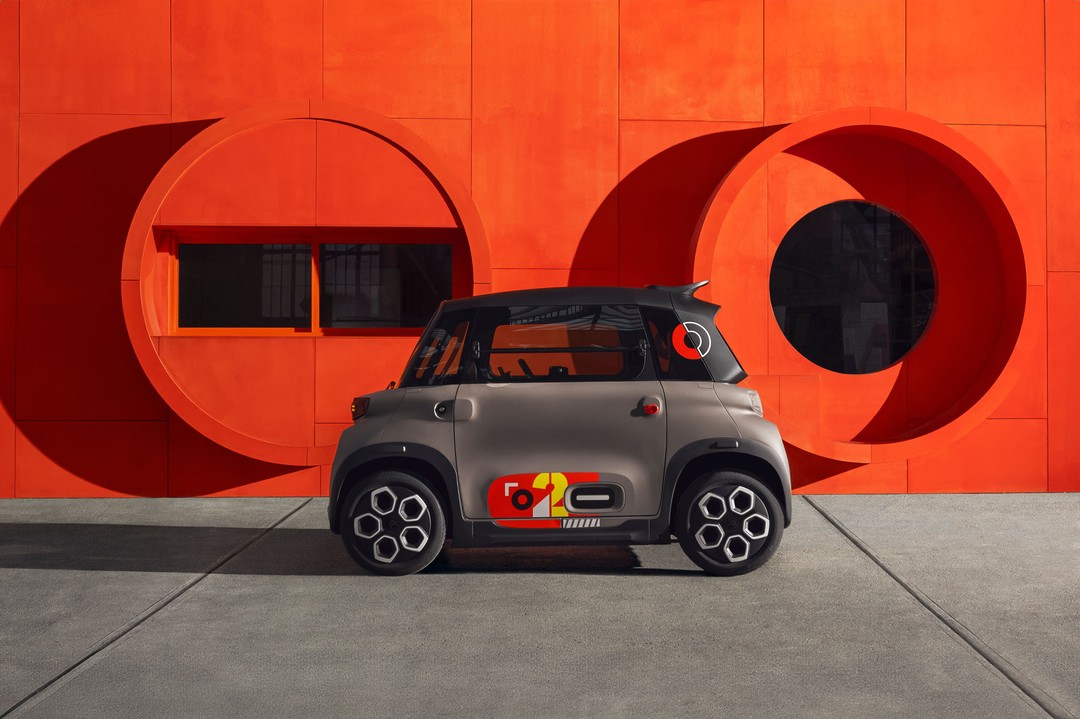 Compact gray Citroen Ami electric car parked in front of an orange wall with circular cutouts, on a gray tiled surface.