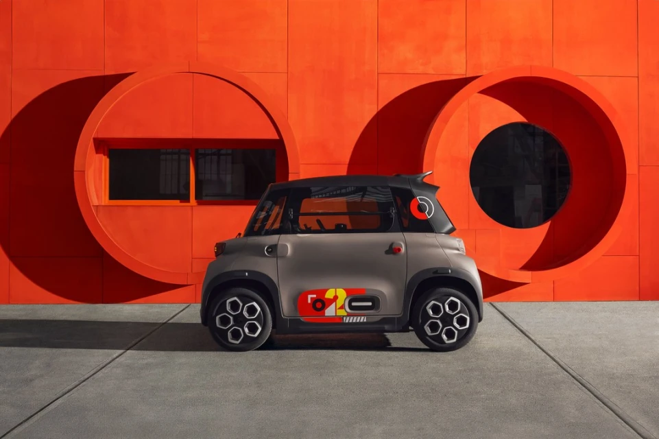 Compact gray Citroen Ami electric car parked in front of an orange wall with circular cutouts, on a gray tiled surface. 