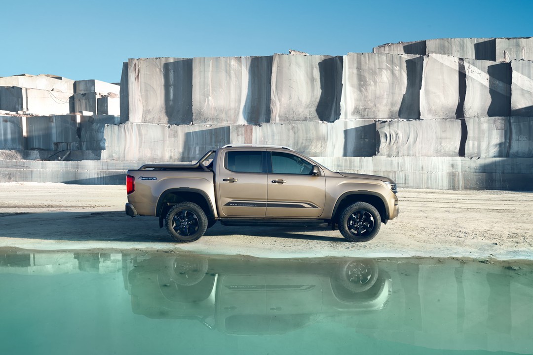 A bronze pickup truck parked next to a still body of water with a quarry wall backdrop and clear blue sky above on a sunny day.