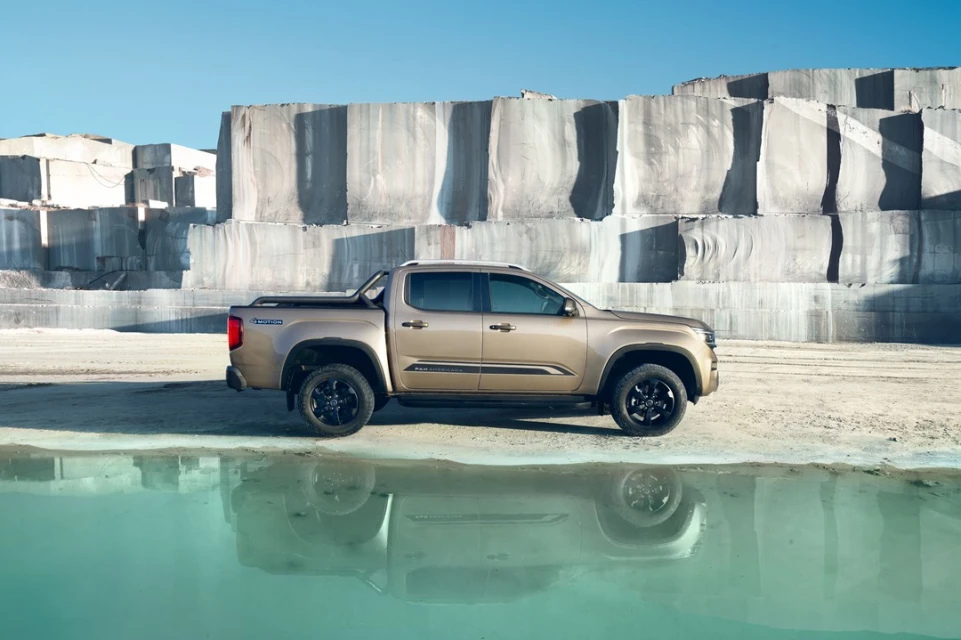 A bronze pickup truck parked next to a still body of water with a quarry wall backdrop and clear blue sky above on a sunny day.