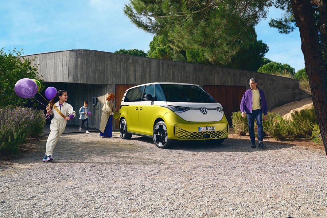 Volkswagen ID. Buzz electric van parked outdoors with a family of four; a girl runs toward the camera with balloons.