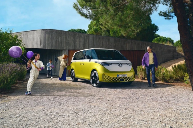 Volkswagen ID. Buzz electric van parked outdoors with a family of four; a girl runs toward the camera with balloons.