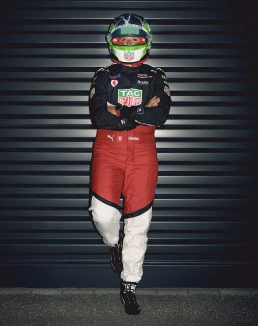Full-shot of a race car driver leaning against a wall, wearing a racing suit and helmet with arms crossed in an indoor setting.