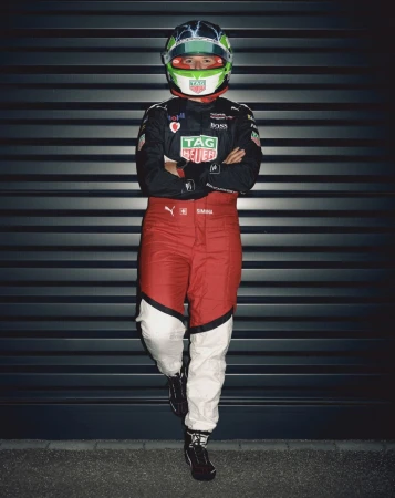 Full-shot of a race car driver leaning against a wall, wearing a racing suit and helmet with arms crossed in an indoor setting.
