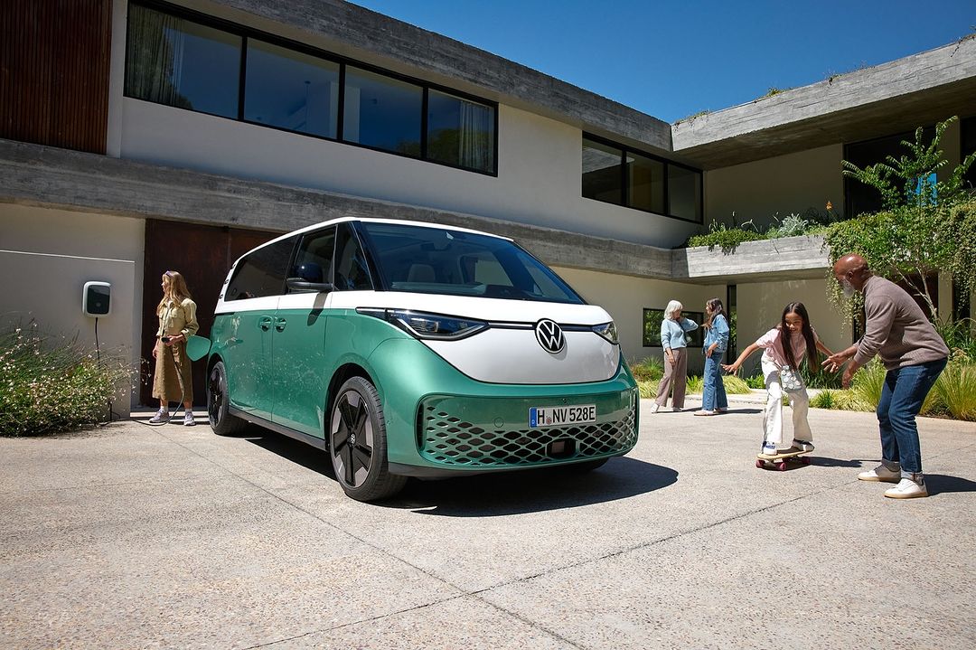 Mint green and white VW ID. Buzz minivan parked outside a modern house, with family members gathered around. Bright, sunny day.