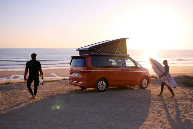 Orange camper van parked on a sandy beach at sunset with two people in wetsuits holding surfboards walking nearby.