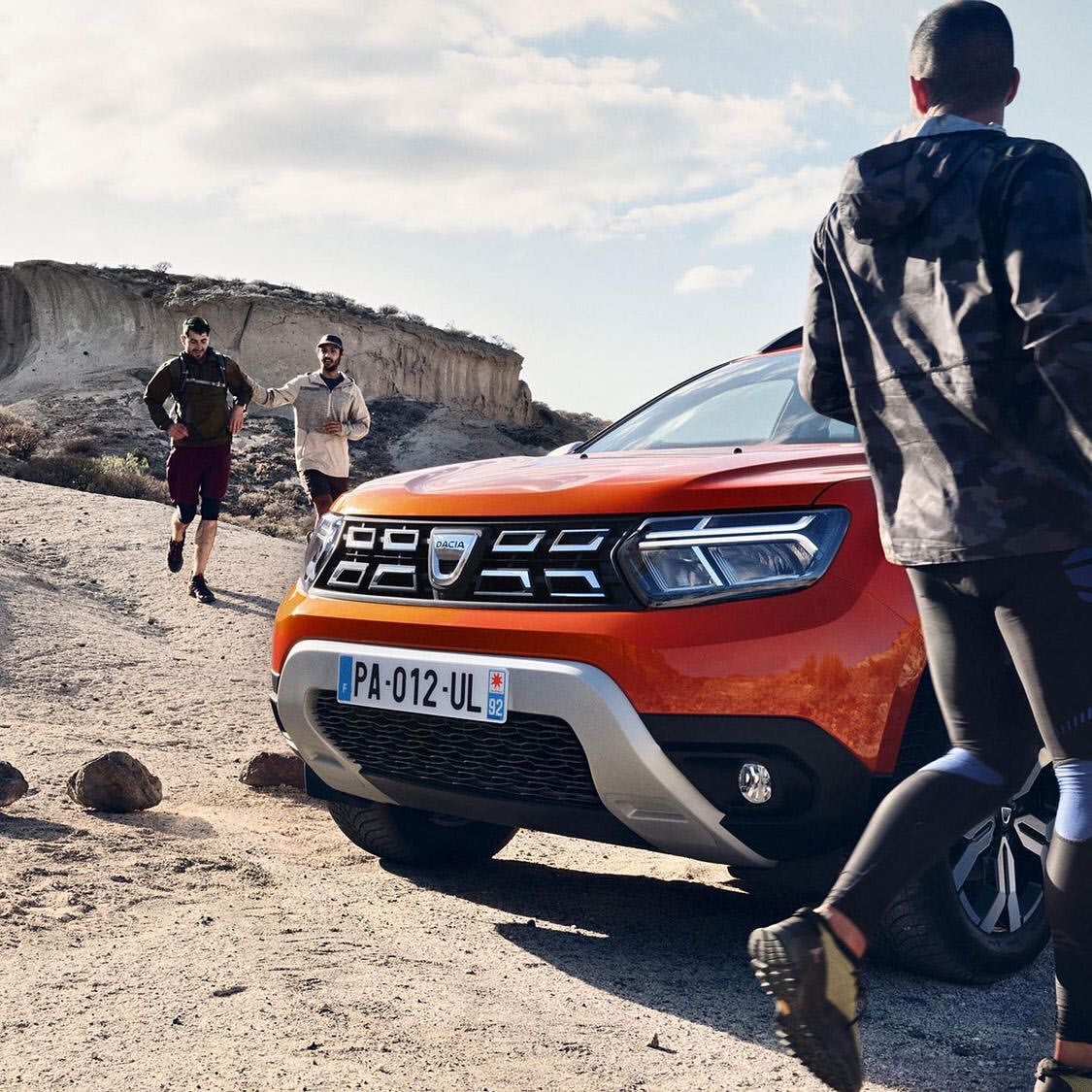 Orange Dacia Duster SUV on a dirt road with two men running in the background and one man blurred in the foreground, daytime.