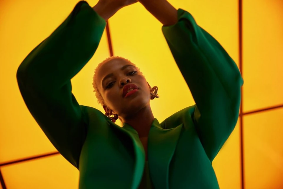 Portrait of a Black female model in a green blazer, posing against a yellow geometric background with raised hands.