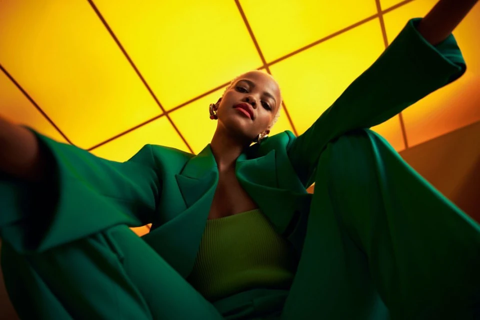 Stylish woman in a green suit poses against a yellow geometric background, shot from a low angle highlighting fashion and modern aesthetics.