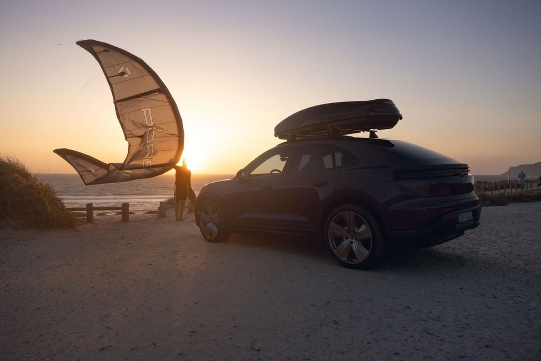 Black SUV with a roof box parked on a sandy beach at sunset with a person holding a kite in the background by the ocean.