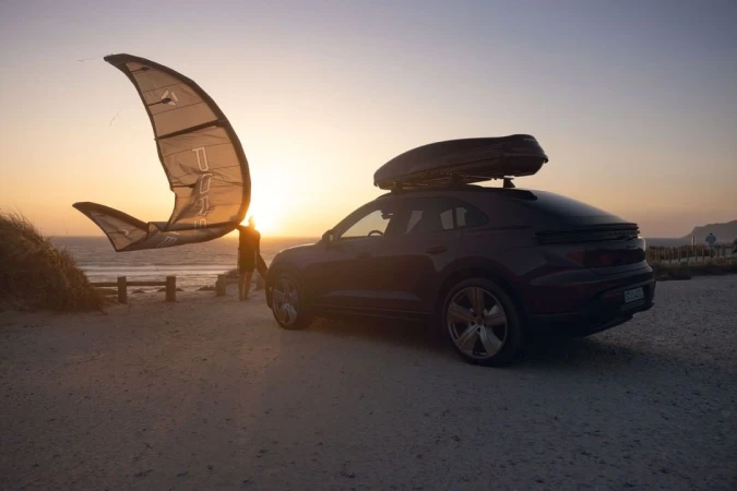 Black SUV with a roof box parked on a sandy beach at sunset with a person holding a kite in the background by the ocean.