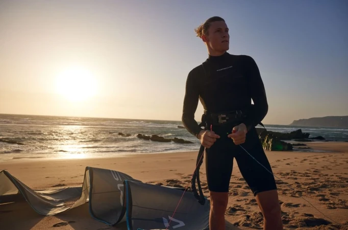 Adult male kitesurfer stands on sandy beach in black wetsuit holding kite lines, sun behind him, with ocean and shoreline background.