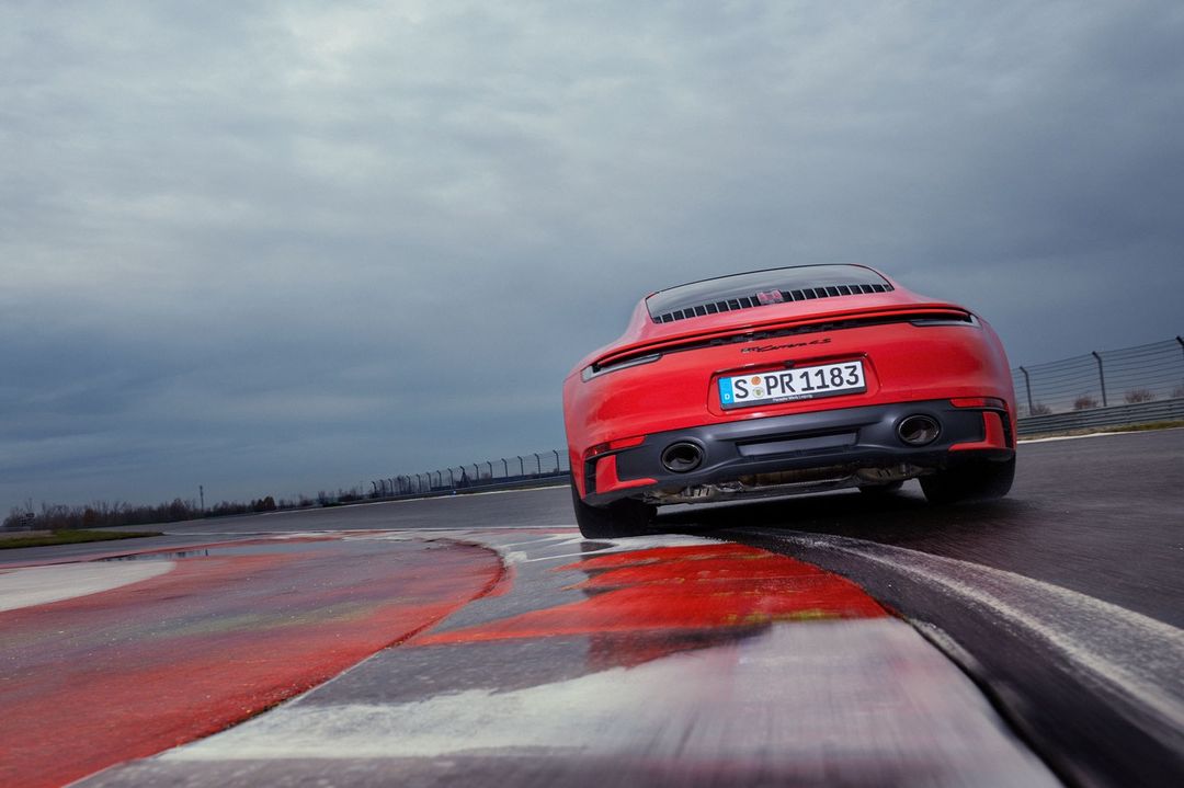 Rear view of a red Porsche 911 Carrera S on a race track with a cloudy sky. Vehicle is angled on the track with curbing visible.