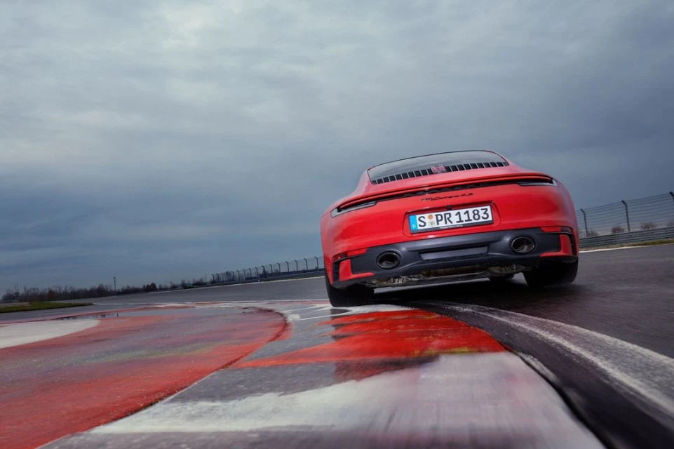 Rear view of a red Porsche 911 Carrera S on a race track with a cloudy sky. Vehicle is angled on the track with curbing visible.