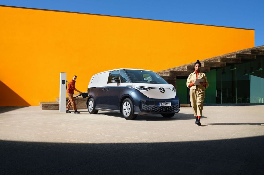 Volkswagen ID. Buzz cargo van charging with two people, one walking and the other holding the charger against an orange building.