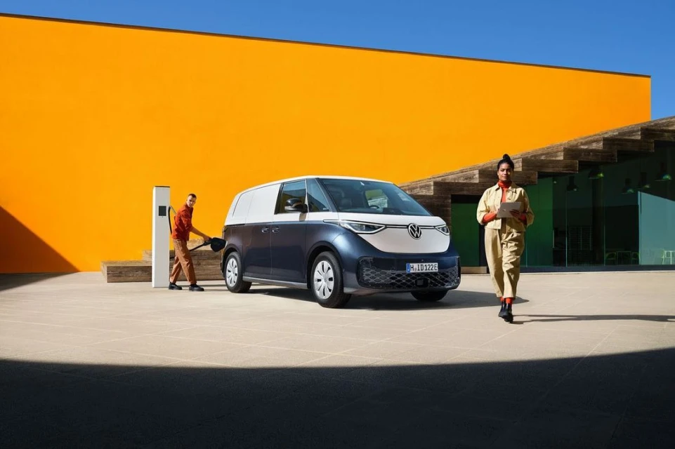 Volkswagen ID. Buzz cargo van charging with two people, one walking and the other holding the charger against an orange building.