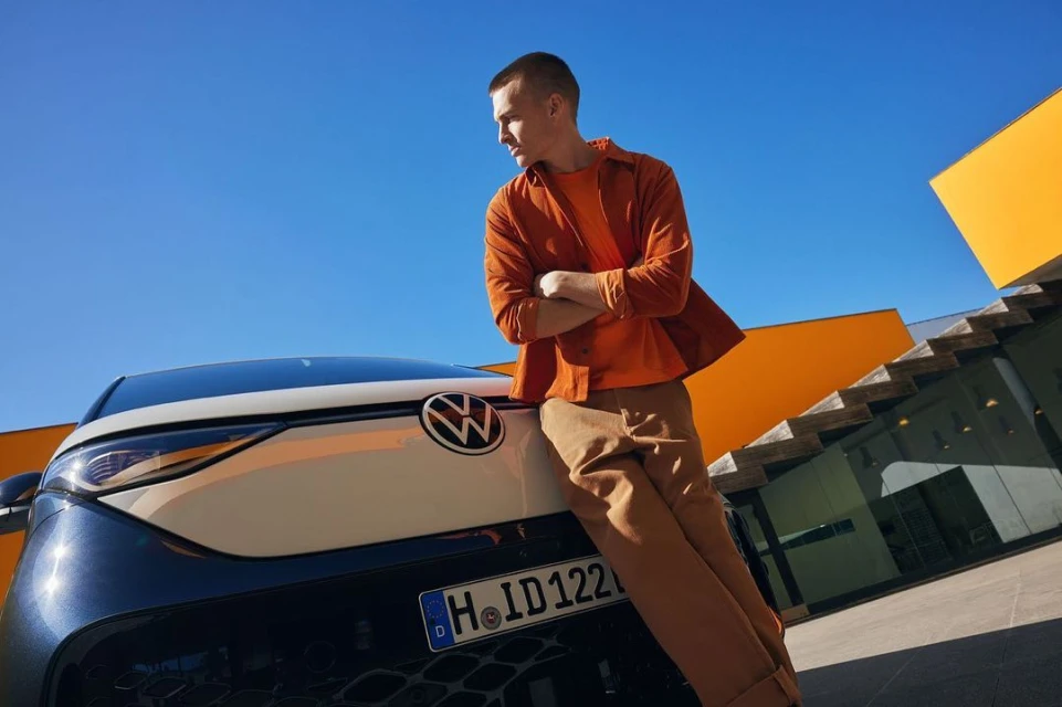 Adult male leaning against a two-tone white and navy blue Volkswagen car outdoors under a clear sky next to an orange building.