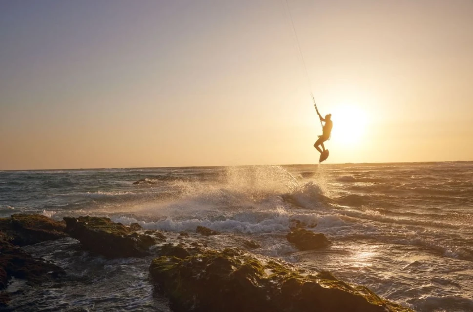 Silhouette of kitesurfer catching air over ocean waves at sunset. Rocky shoreline on left. Water sprays in foreground. Golden sunlight.