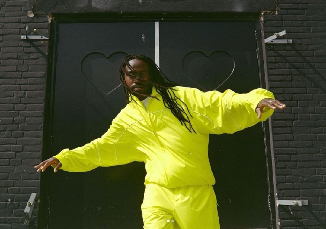 Man in neon yellow tracksuit strikes dynamic pose in front of black brick wall and heart-decorated door, fashion-forward statement.