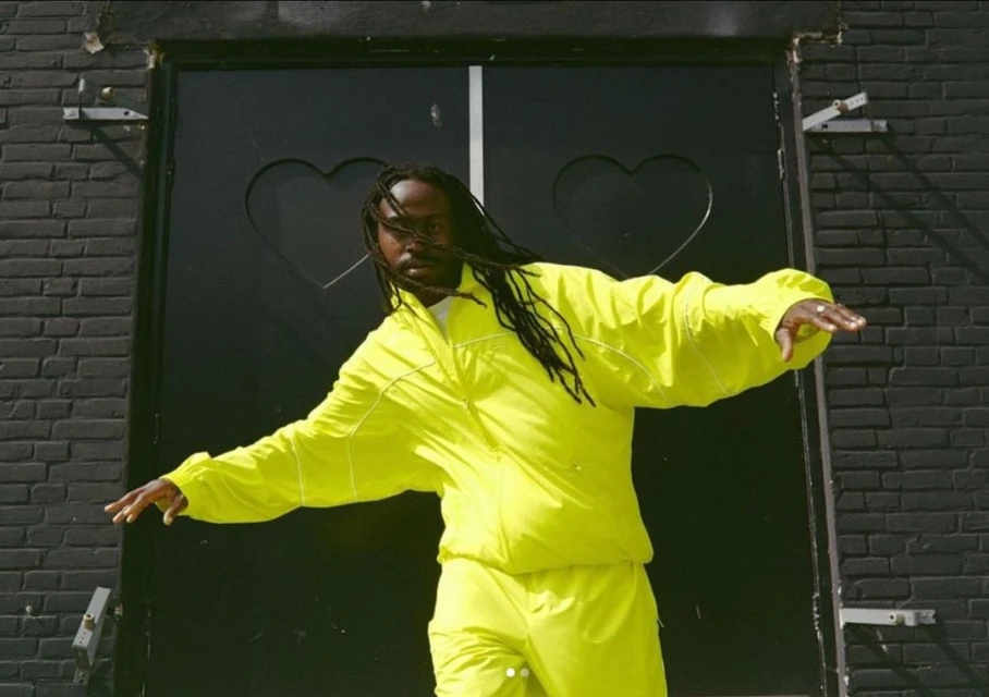 Man in neon yellow tracksuit strikes dynamic pose in front of black brick wall and heart-decorated door, fashion-forward statement.