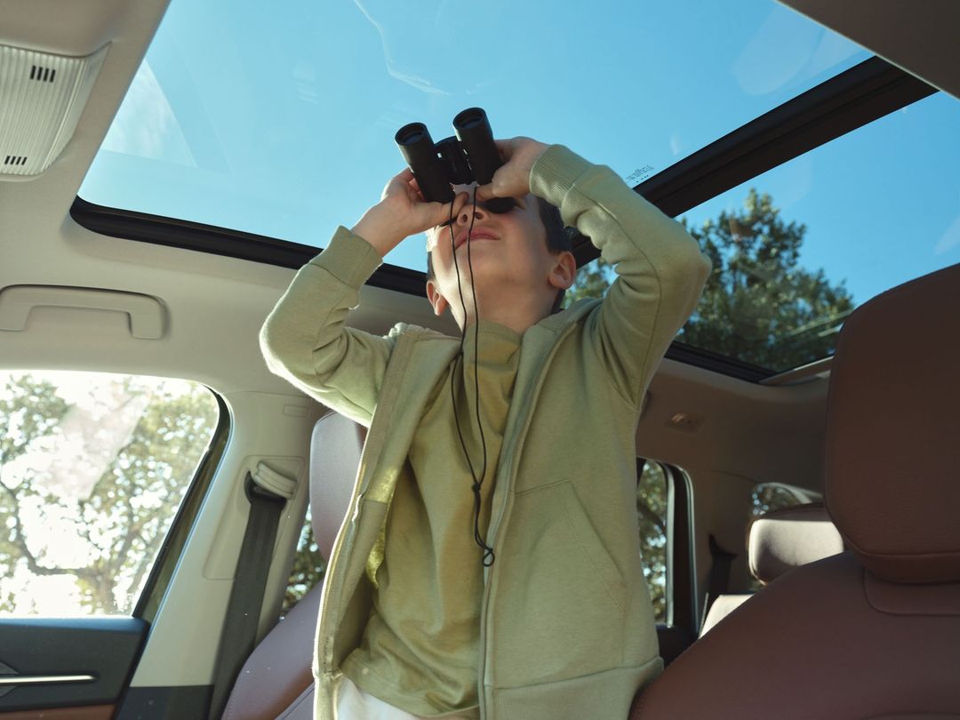 A child sits inside a car with an open sunroof, looking through binoculars up at the sky with trees visible in the background.