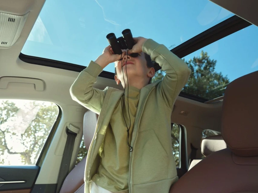 A child sits inside a car with an open sunroof, looking through binoculars up at the sky with trees visible in the background.