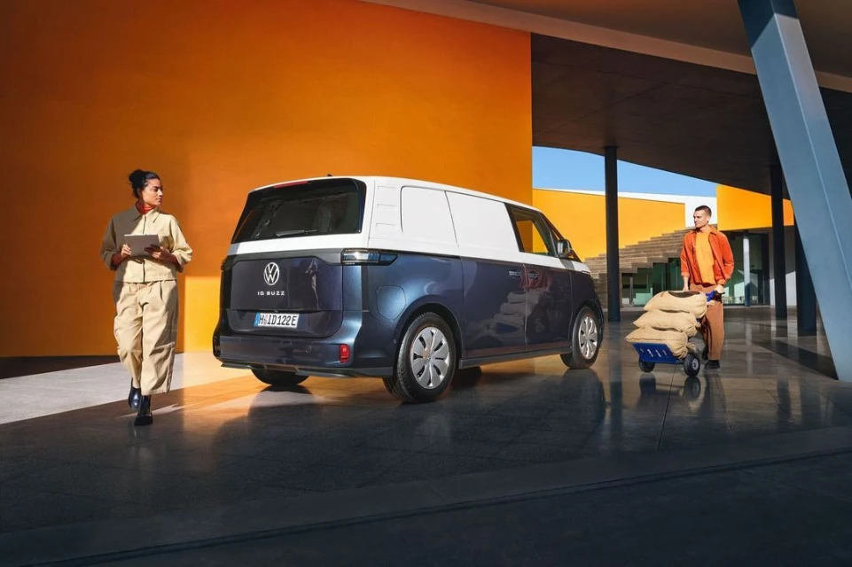 Rear view of a two-tone blue and white Volkswagen ID. Buzz cargo van with a woman holding a tablet and a man with a hand truck.