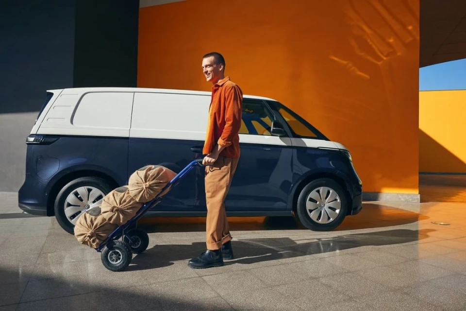 Delivery man pushing dolly with burlap sacks in front of a white and blue Volkswagen ID. Buzz electric cargo van outdoors.