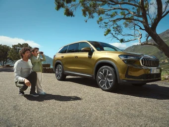 Man and young child looking through binoculars next to a parked gold SUV on an asphalt road near trees and water.