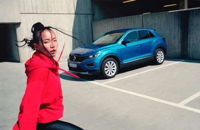 Woman in vibrant red hoodie standing outdoors near a parked blue SUV against a concrete wall