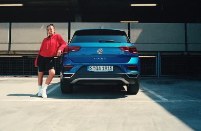 Woman in red hoodie leaning against the back of a blue Volkswagen T-Roc in a concrete parking structure