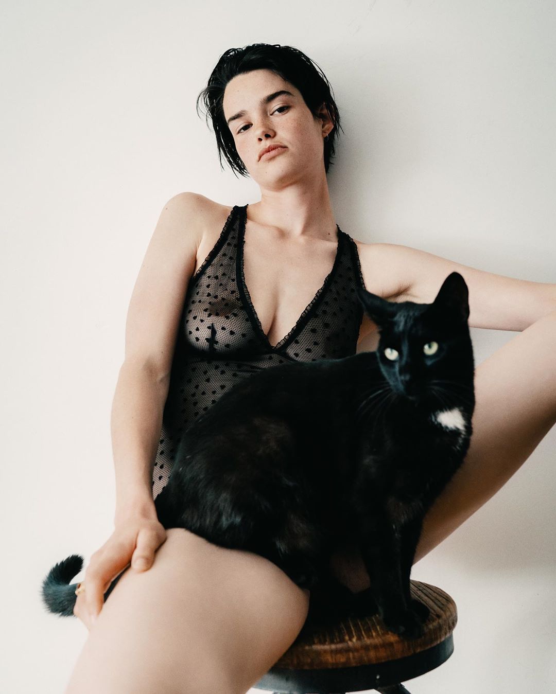 Woman in a patterned bodysuit sitting on a stool, with a black cat perched on her lap. Clean indoor backdrop and medium camera framing.