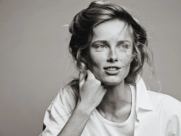 Monochrome portrait of a woman with freckles wearing a white t-shirt, resting her hand on her neck against a plain grey background.