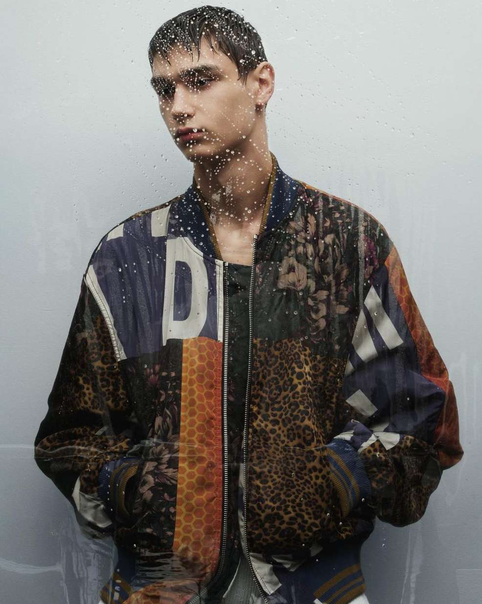 Male fashion model poses indoors in a vibrant, patterned bomber jacket. Water droplets on the foreground suggest a recent rainfall.