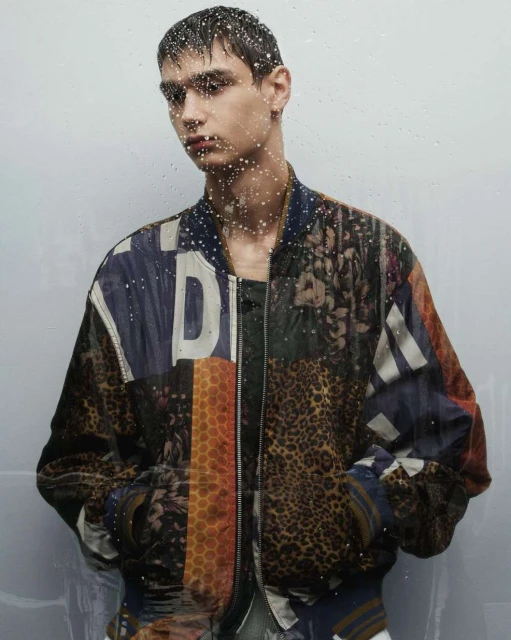 Male fashion model poses indoors in a vibrant, patterned bomber jacket. Water droplets on the foreground suggest a recent rainfall.