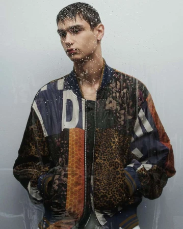 Male fashion model poses indoors in a vibrant, patterned bomber jacket. Water droplets on the foreground suggest a recent rainfall.