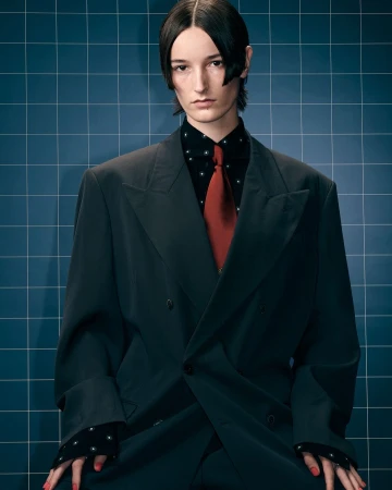 Person wearing a dark oversized suit with a red tie poses against a blue tiled wall in a studio fashion portrait. 
