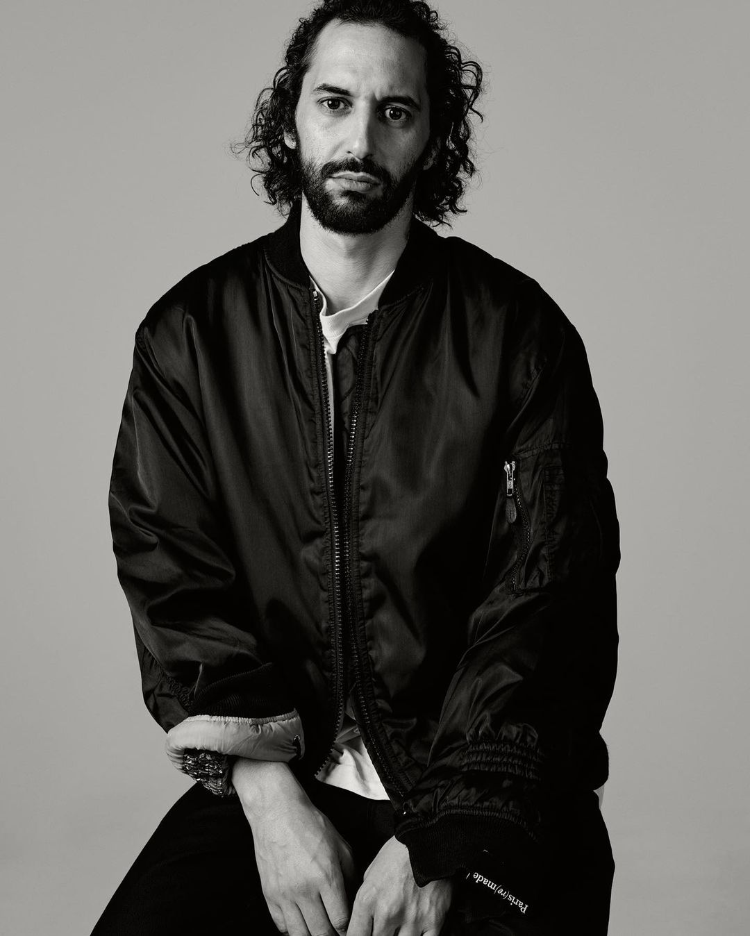 Monochrome portrait of adult male with curly hair and beard in a bomber jacket, seated indoors against a neutral grey background.
