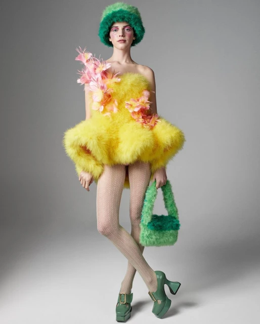 A model poses in a fluffy yellow mini-dress and green accessories, including a hat, purse and chunky platform shoes against grey.