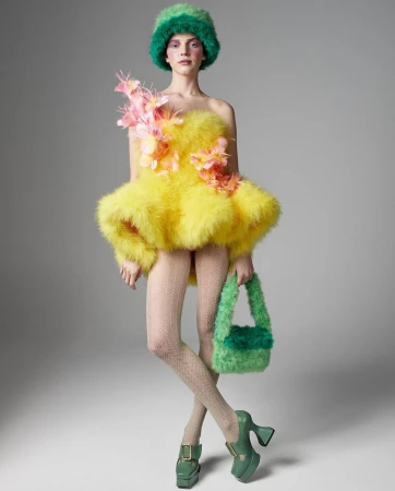 A model poses in a fluffy yellow mini-dress and green accessories, including a hat, purse and chunky platform shoes against grey.
