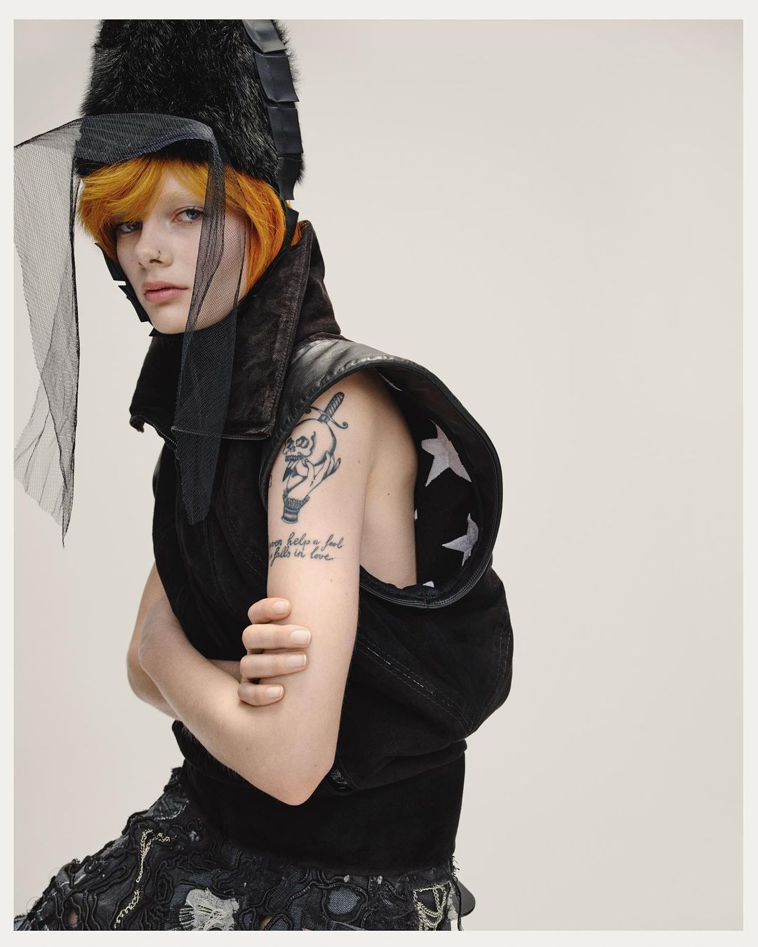 A pale-skinned person with orange hair, dark vest, and hat with netting poses indoors against a plain background, wearing edgy fashion.