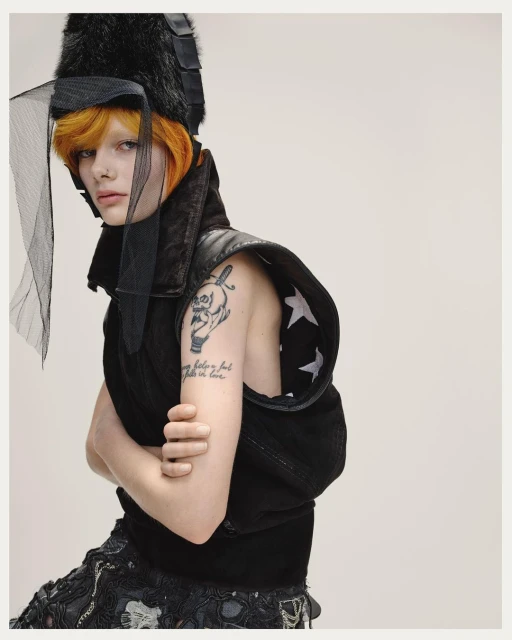 A pale-skinned person with orange hair, dark vest, and hat with netting poses indoors against a plain background, wearing edgy fashion.