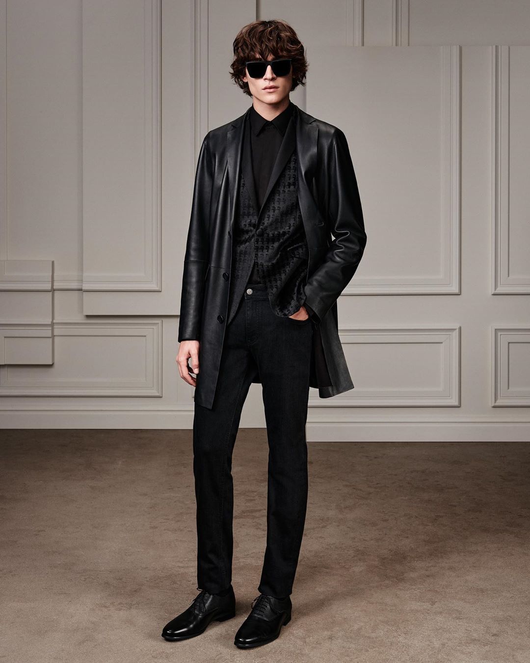 A stylish adult male stands indoors wearing an all-black outfit with a black leather trench coat and sunglasses, posing in a studio.