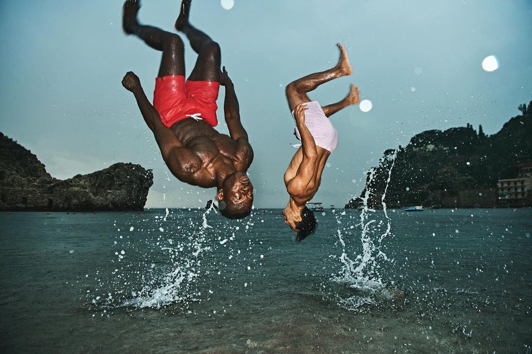Two men in mid-air performing backflips over ocean with rocky coastline in background; water splashing up from below.