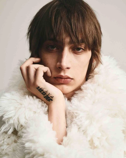 Close-up studio shot of a young adult with shoulder-length brown hair wearing a fluffy white jacket, gazing directly at the camera with hand near face.