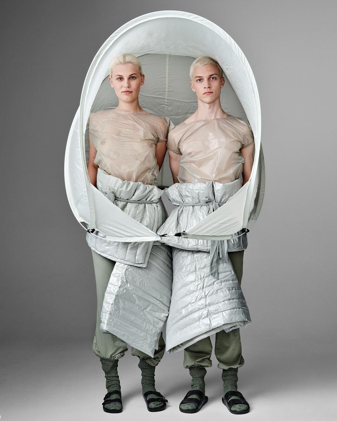 Two fair-skinned adults in a studio pose, wrapped in a large white oval frame and wearing neutral tops, puffer shorts, socks, and sandals.