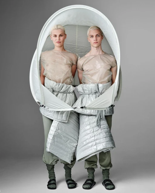 Two fair-skinned adults in a studio pose, wrapped in a large white oval frame and wearing neutral tops, puffer shorts, socks, and sandals.