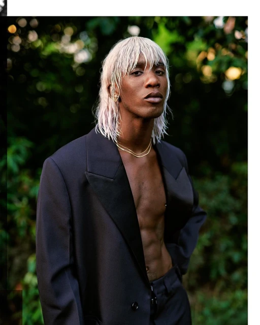 Stylish man with blonde mullet and dark suit poses in an outdoor setting, showing his toned torso and wearing a gold necklace.