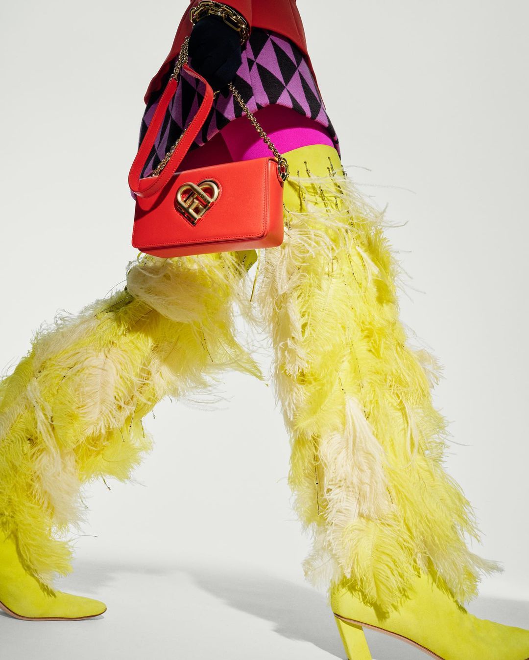 Fashion editorial featuring yellow feather-embellished pants and boots paired with a bright red heart-shaped handbag on a white background.
